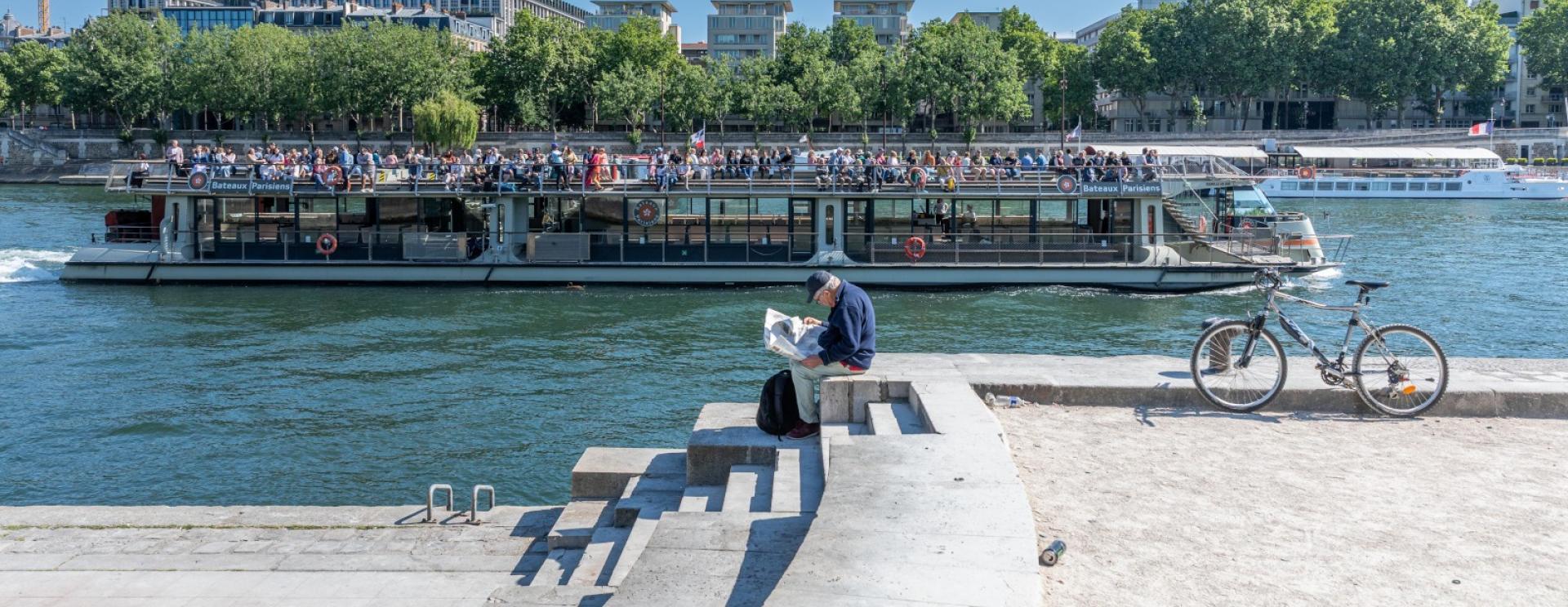 Scène de vie sur les quais de Seine à Paris sur le quai Saint Bernard. Un bateau de croissière navigue sur l'eau pendant qu'au premier plan un vieil homme lit son journal assis sur des marches.